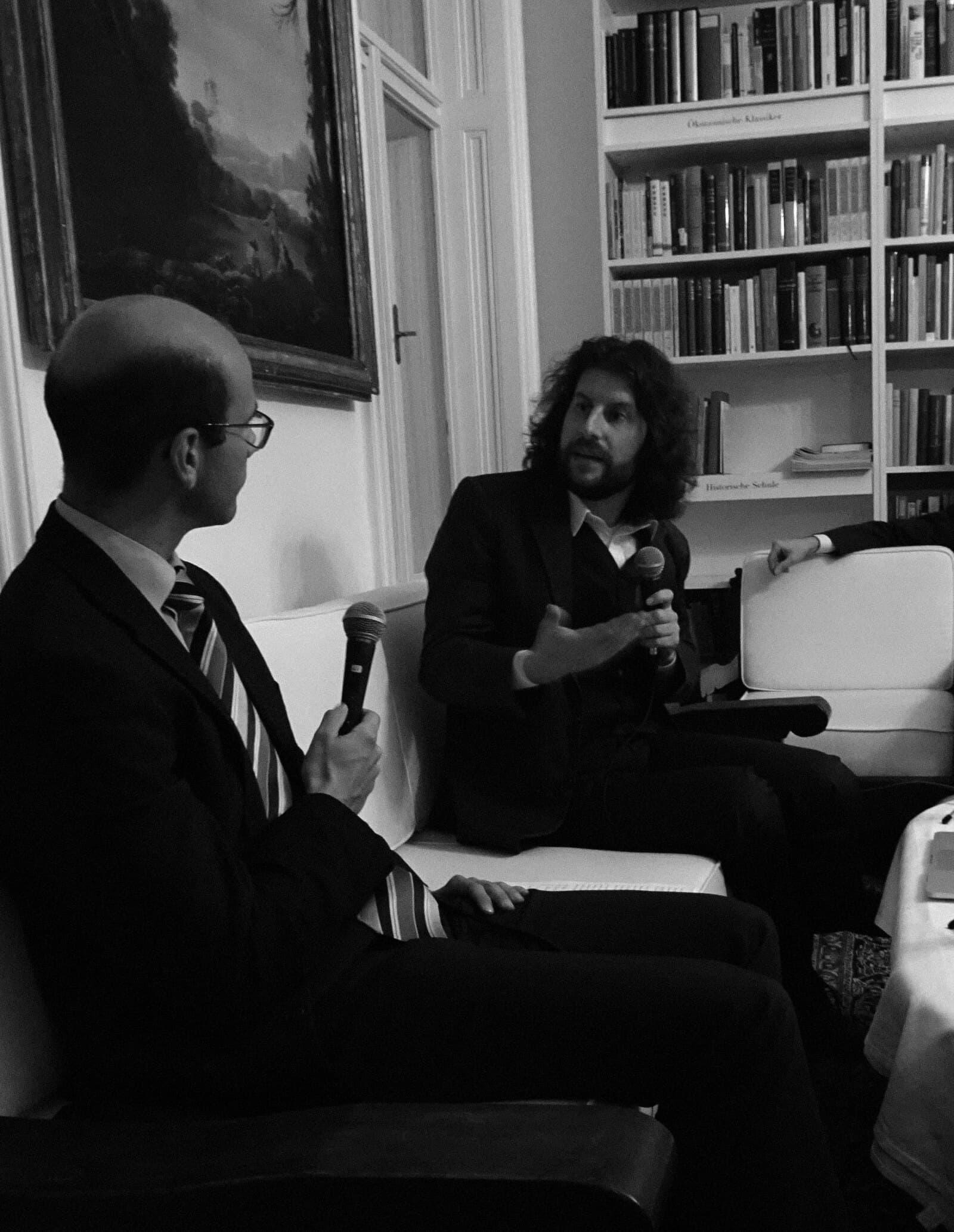 Rahim Taghizadegan im Gespräch mit einem Gast, beide mit Mikrofon, vor den Regalen der Wiener Bibliothek