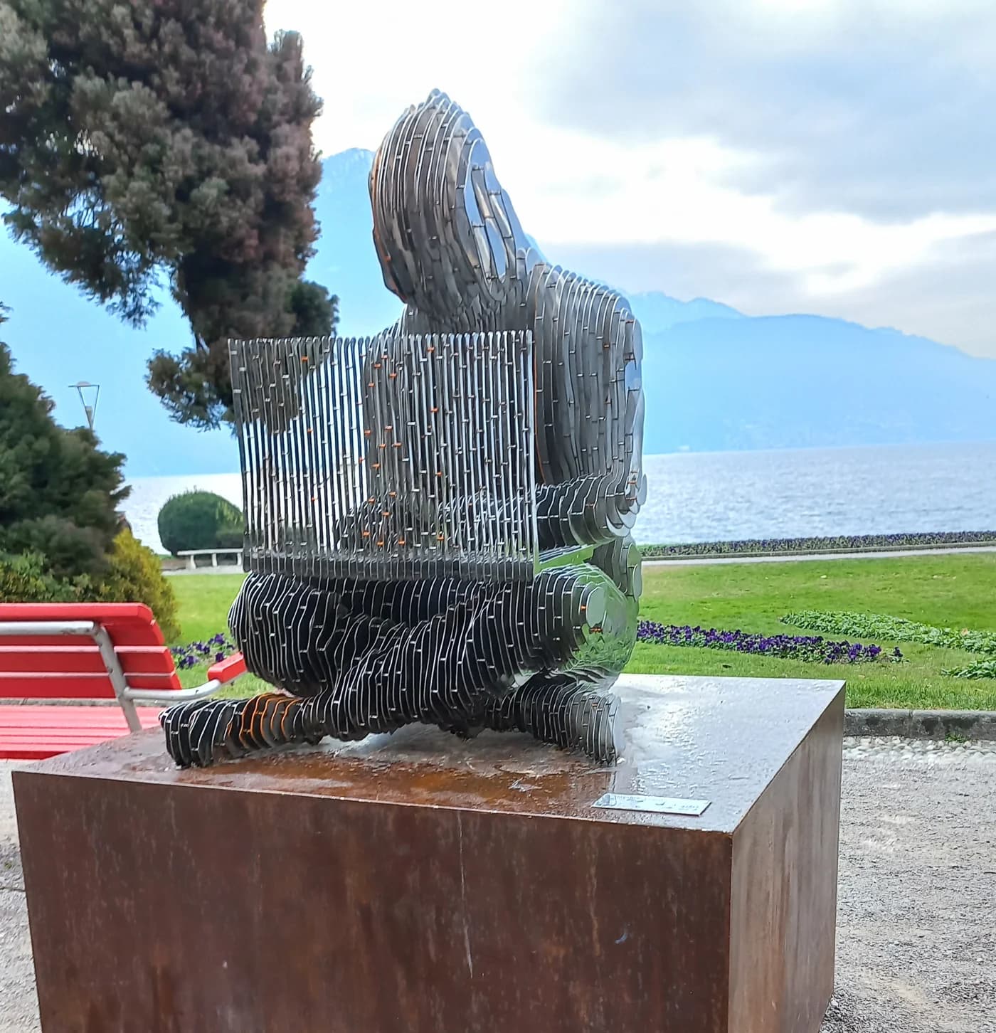La estatua de Satoshi en Lugano: una figura sentada con un portátil, hecha de láminas de acero inoxidable, sobre un pedestal oxidado, con el lago de Lugano al fondo.